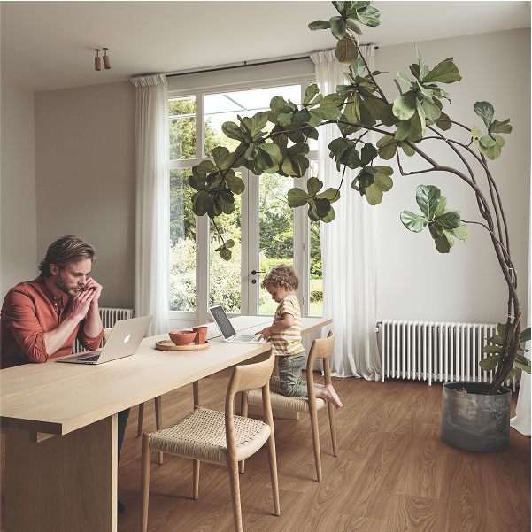 A man and a child work on laptops at a wooden dining table in a bright room with large windows, white curtains, TORLYS flooring, and a tall indoor plant in the corner.