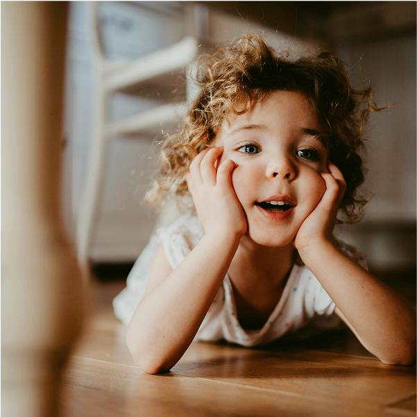 A young child with curly hair lies on TORLYS flooring, propping their face in their hands and smiling, as a soft, warm light illuminates the inviting scene.