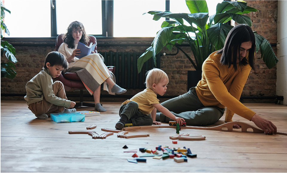 Two children play with wooden train tracks and blocks on smart floors while an adult helps them. Another adult sits in the background on a chair, reading a book. Large green plants and a window are in the background.