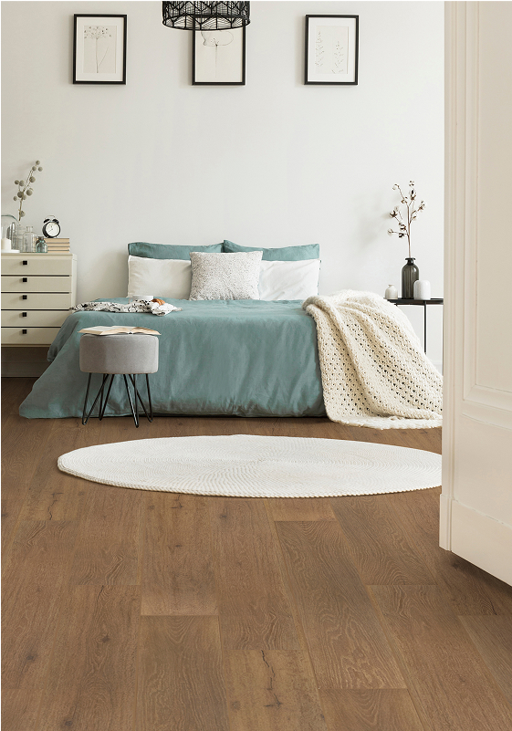 A cozy bedroom with a teal bed, white blanket, round white rug atop smart floors, minimalist decor, a small side table, and framed art on the white wall.