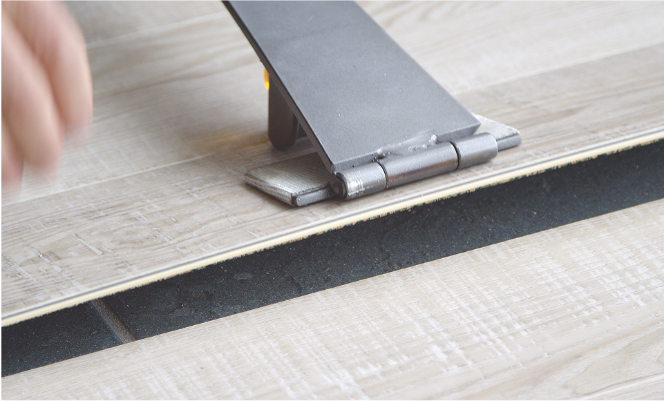 A close-up of a tool being used to install or adjust smart floors with light-colored wooden or laminate finish, a hand visible on the left side of the image.