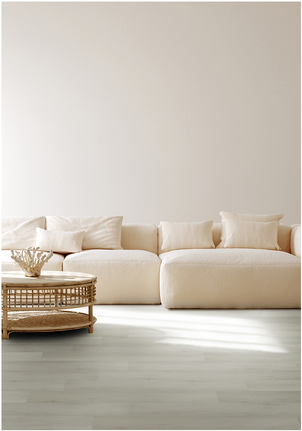 A minimal living room with a large cream-colored sectional sofa, white pillows, a round wicker coffee table, and light wood smart floors, set against a plain off-white wall.