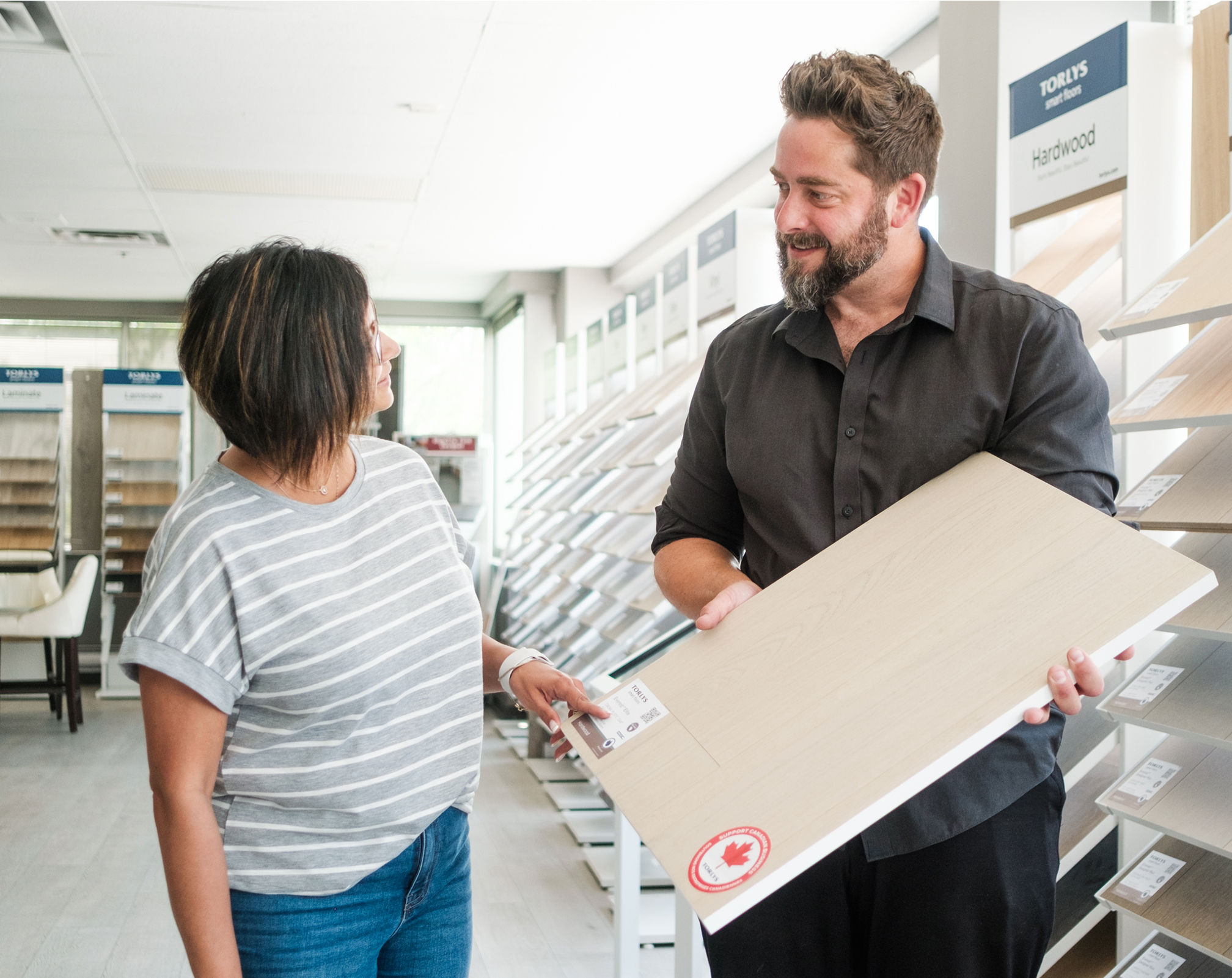 A woman and a man stand in a flooring showroom. The man holds a large sample of light-colored wood flooring while the woman points at a label on it. Both appear to be discussing the product.