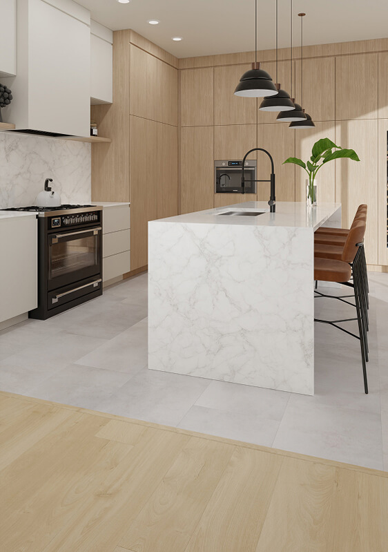 Modern kitchen featuring TORLYS flooring, a white marble island with three brown barstools, black faucet, hanging black pendant lights, light wood cabinets, built-in oven, and a large green plant as an accent.