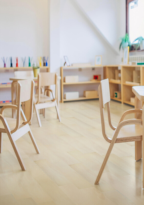 A bright, tidy classroom with wooden chairs and tables, TORLYS flooring underfoot, open shelves holding books and art supplies, and natural light streaming through a window with a potted plant on the sill.