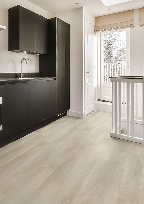 A modern kitchen with matte black cabinets, TORLYS flooring in light wood tones, a white door with glass panels, and a staircase railing to another level. Natural light pours in through a large window and door.