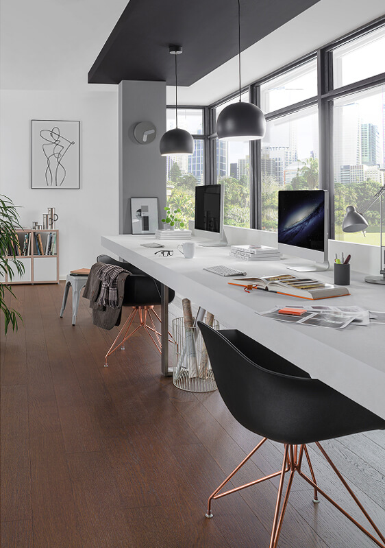 Modern office space with large windows, two black chairs at a long white desk, TORLYS flooring, computers, books, plants, pendant lights, and a cityscape view. Minimalist decor includes framed art and shelves.