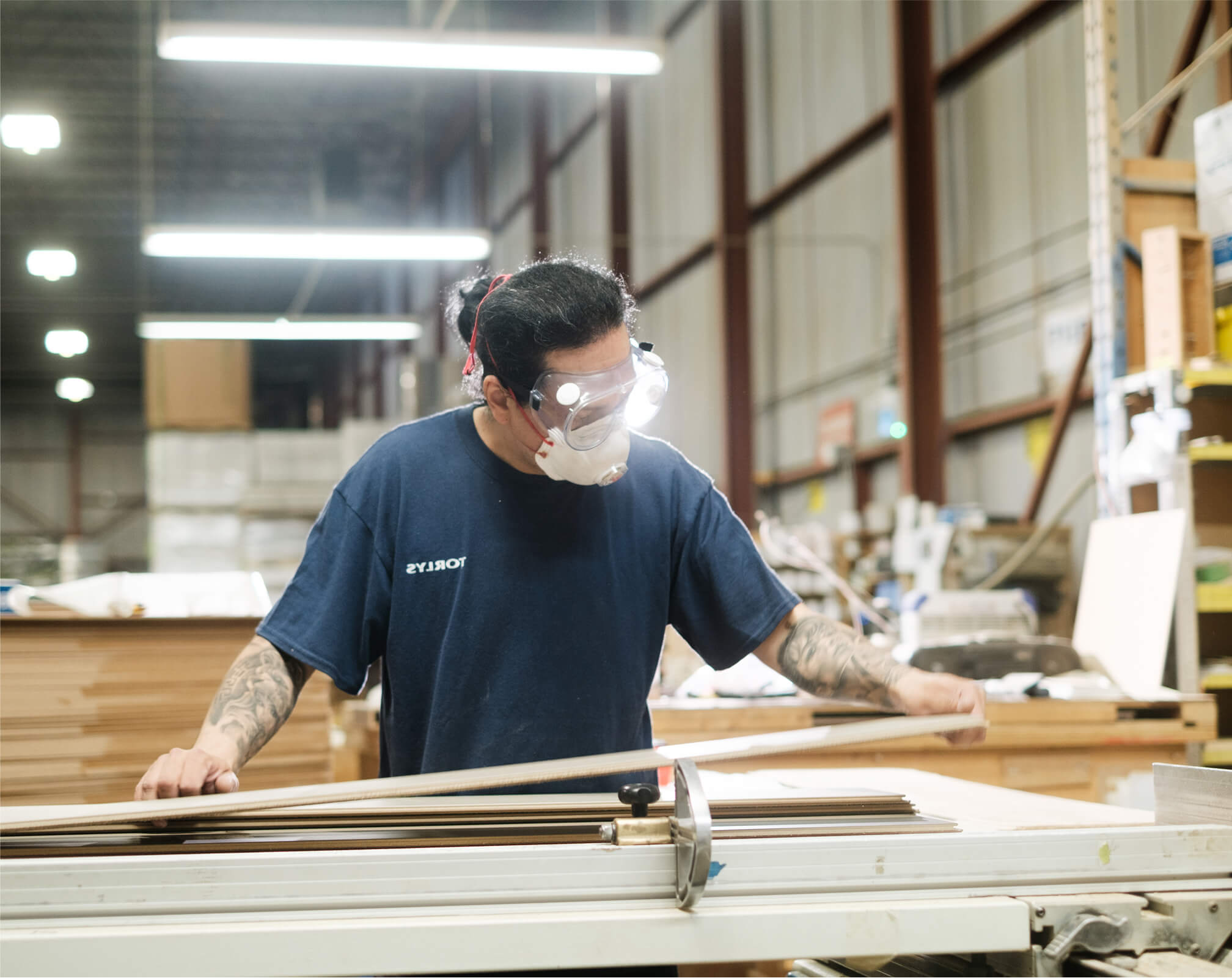 A person wearing safety goggles, a face mask, and a navy blue shirt operates a table saw on smart floors in a well-lit industrial workshop with shelves and stacked materials in the background.
